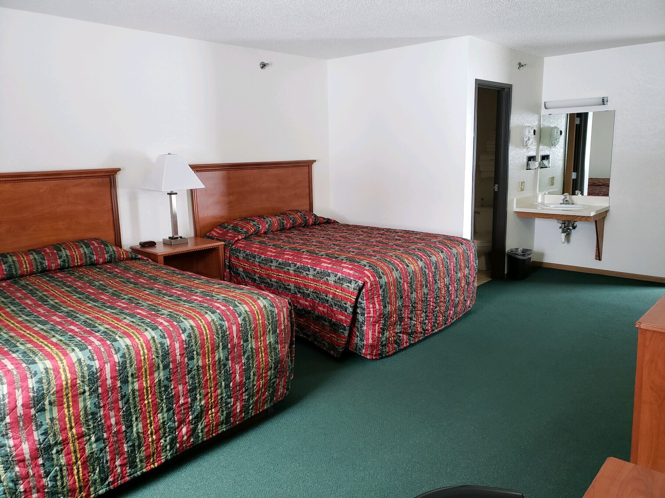 A hotel room with two beds, a nightstand, a lamp, a sink, and a mirror.