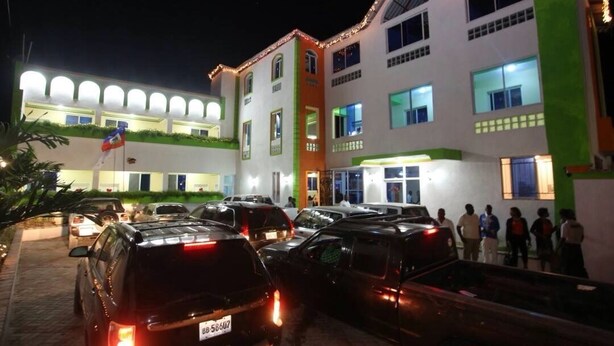 Un hôtel à étages, un parking où sont garées plusieurs voitures, et un groupe de personnes près de l’entrée.