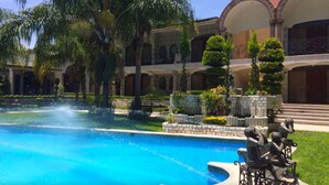 A pool with a fountain, surrounded by palm trees and a building with arches and columns.