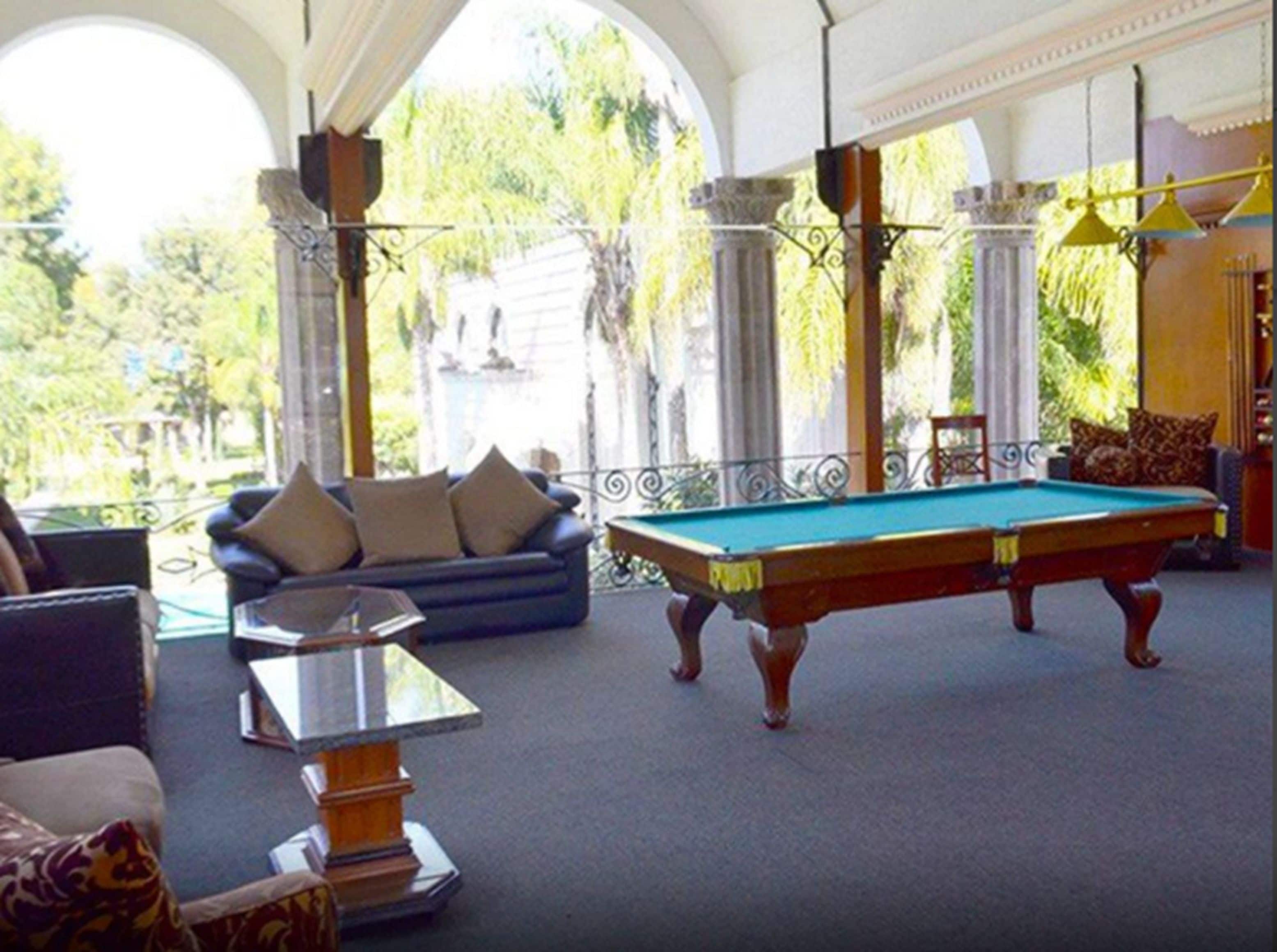 A pool table in a room with a sofa and a table, large windows with a view of greenery outside.