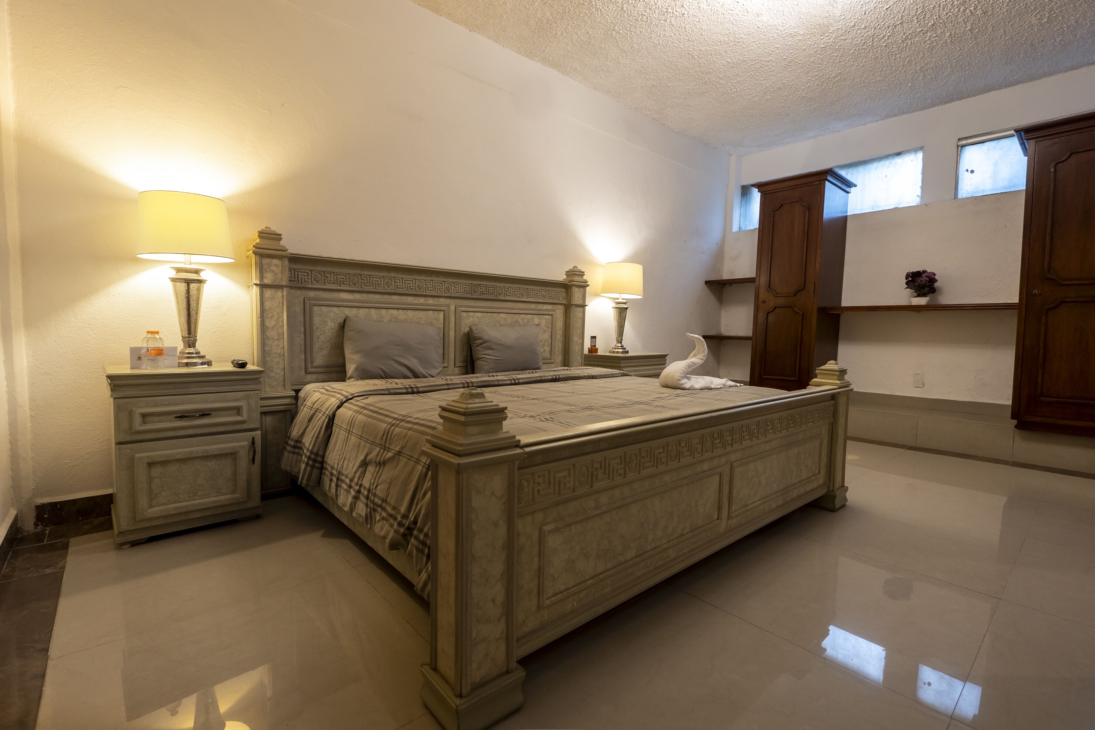 A bedroom with a large bed, two bedside tables with lamps, a nightstand, a dresser, and a window with a view of the sky.
