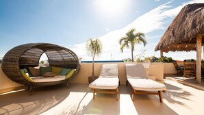 A sunlit outdoor seating area with a woven chair and two lounge chairs, a thatched-roof structure, and palm trees in the background. - Lovely Kingsize-bed Studio, amazing rooftop with pool, best location (unit 203) (Playa del Carmen)
