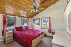 Un dormitorio con techo de madera, cama con colcha roja, escritorio y dos ventanas con cortinas.