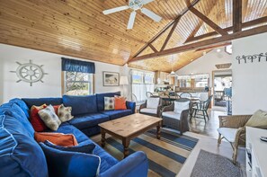 Una sala de estar acogedora con un sofá azul, una mesa de centro de madera y sillas de mimbre. Se ve una cocina al fondo.