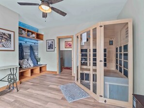 Un pasillo con ventilador de techo, estantes de madera con ropa doblada, una mesita con lámpara y una puerta de madera con paneles de vidrio.