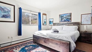 Een slaapkamer met een bed, een raam met gordijnen, een vloerkleed en foto's in lijsten aan de muur.