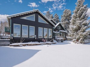 A two-story house with large windows, a covered porch, and snow-covered trees. - Mountain Hideaway — Hot Tub | Sauna | 1Gbps WiFi | 9BR Sleeps 18 + Pets (Leadville)