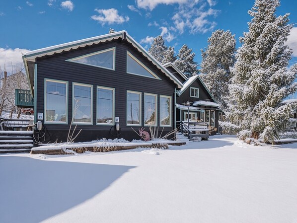 Una casa de dos pisos con amplios ventanales, un porche cubierto y árboles cubiertos de nieve.