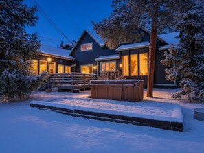Una casa de dos pisos con un porche cubierto y un jacuzzi en un paisaje nevado.