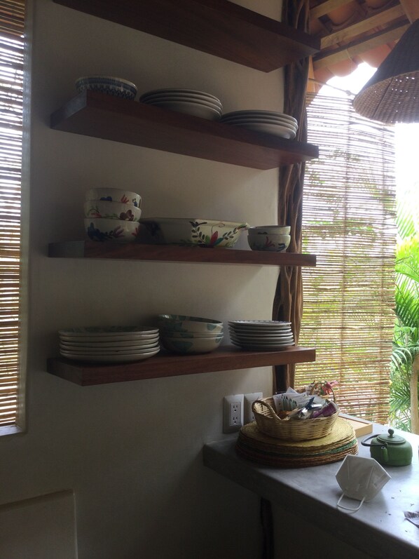 A kitchen with wooden shelves holding plates and bowls. There is a window with blinds, a basket, and a kettle on the counter.