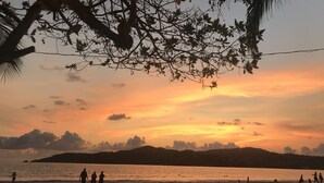 Escena de playa con gente caminando por la orilla, un atardecer en el fondo y un árbol con luces colgantes.