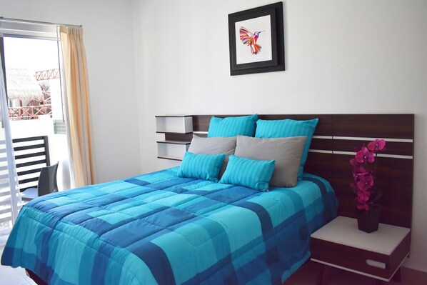 Ein ordentlich bezogenes Bett mit einer blau-taubenfarben karierten Tagesdecke und mehreren Kissen. Darüber an der Wand hängt ein gerahmtes Kunstwerk mit einem bunten Vogel.