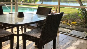 A covered patio area with a glass table and wicker chairs, overlooking a pool and lush greenery. - Fantastic Vacation House on Private Lake in Fort Lauderdale (Fort Lauderdale)