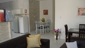 Una sala moderna con un sofá marrón oscuro y una silla blanca, un comedor con mesa y sillas, y una cocina con refrigerador y alacenas.