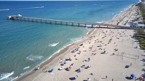 A beach with a long pier extending into the ocean, numerous umbrellas, and a sandy shore. - Modern & Spacious Luxury Apartment – Steps to the Beach (Deerfield Beach)