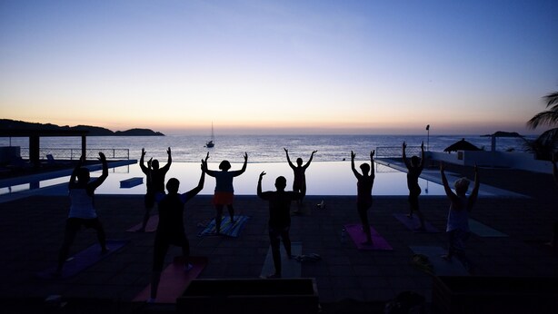 Un grupo de personas practicando yoga sobre esterillas en una playa, con vista al océano y el sol poniéndose en el horizonte.