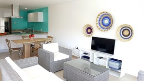Una sala moderna con televisor, muebles de mimbre y una cocina al fondo.