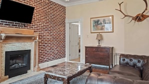 Una sala de estar con chimenea de ladrillo, un televisor de pantalla plana montado encima, un sofá de cuero, una mesa de centro de madera y una decoración de astas de ciervo en la pared.