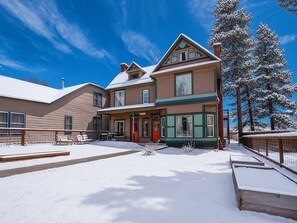 A two-story building with a snow-covered roof and a red door, surrounded by snow and trees. - Governor's Mansion Main — 4BR Sleeps 8, Pool Table, Sauna, Walk to Main St (Leadville)