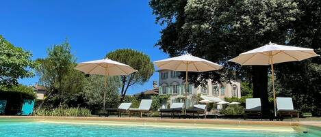 Un área de piscina con sillones y sombrillas, una piscina azul cristalina y un gran árbol al fondo.