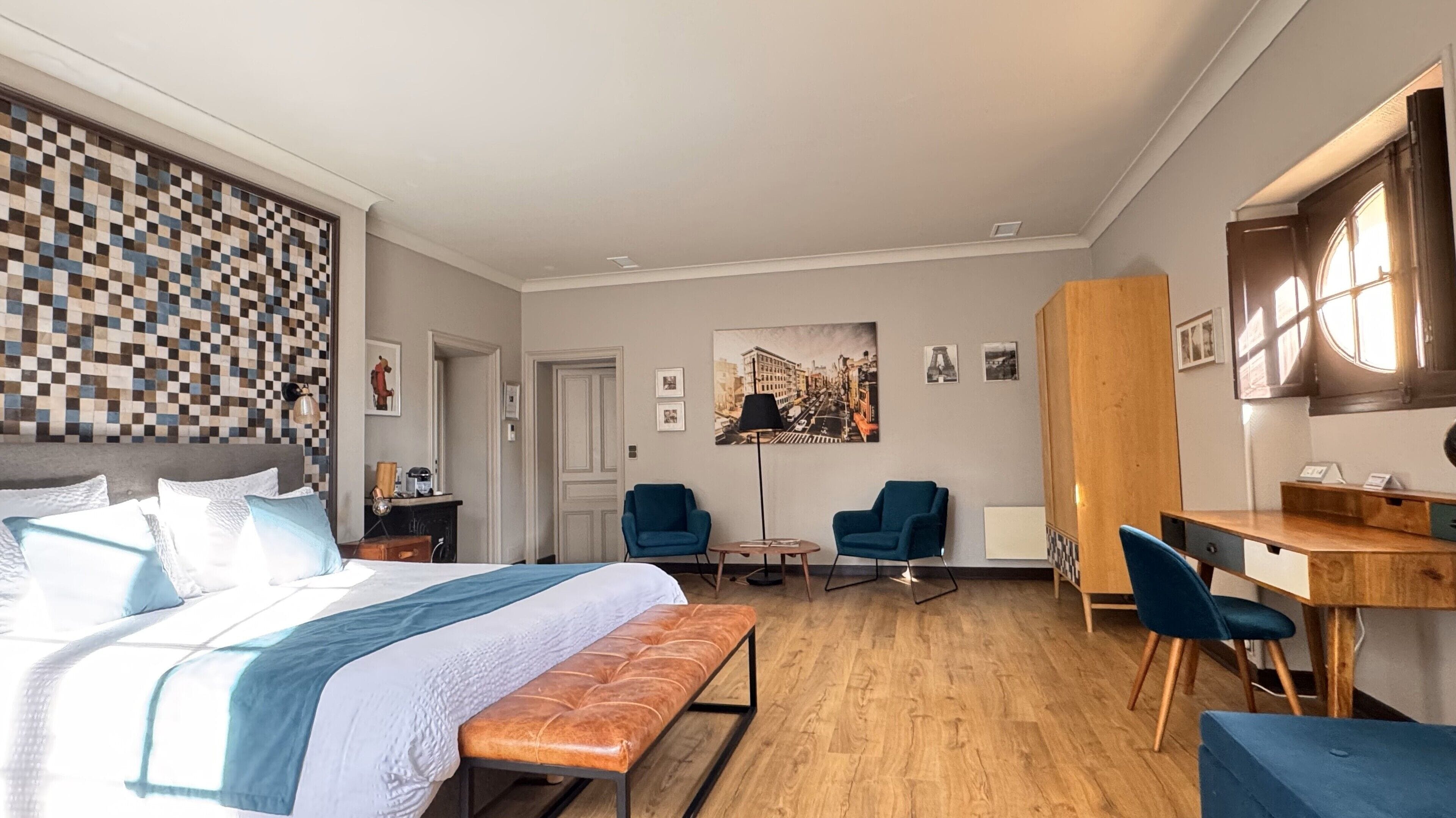 A modern hotel room with a large bed, a desk, two chairs, a bench, and a cityscape painting on the wall.