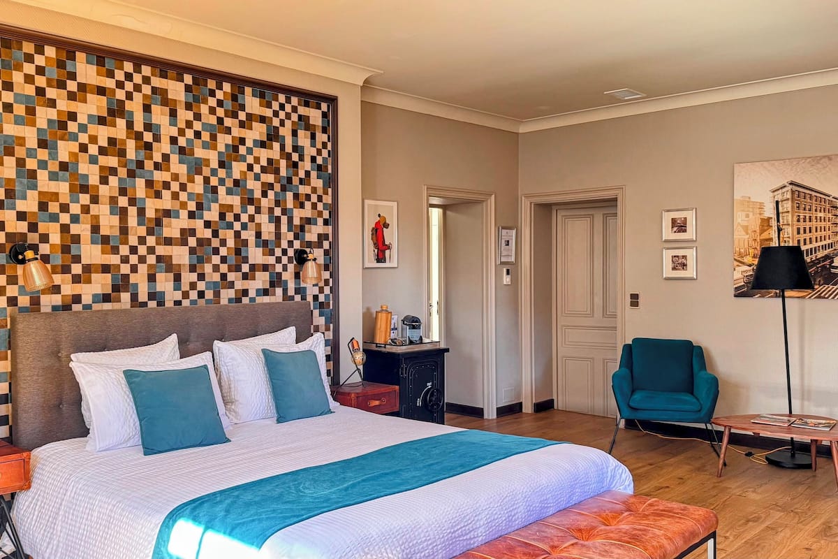 A hotel room with a large bed, a checkered wall, a desk with a lamp, a chair, and a bookshelf.