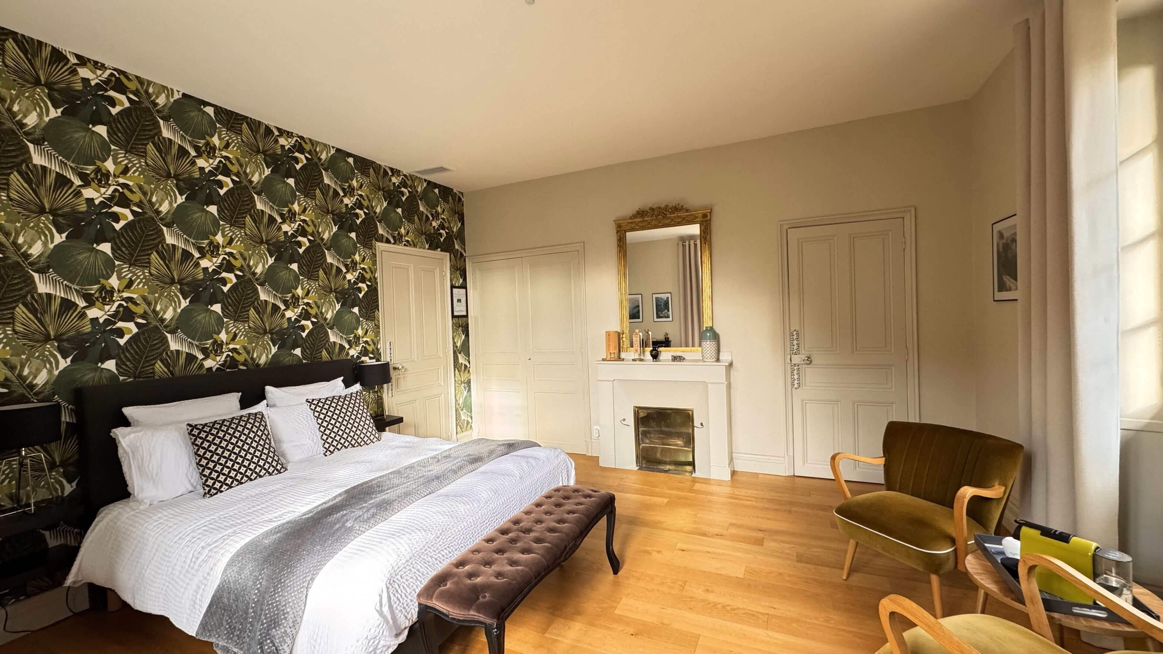 A bedroom with a bed, a bench, a chair, a fireplace, and a mirror.