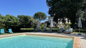 A swimming pool with lounge chairs and umbrellas, surrounded by greenery and a building in the background. - Maison d'hôtes Le Parc (Labruguiere)