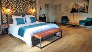 A modern hotel room with a large bed, a bench, two armchairs, a wall-mounted light, and a cityscape painting. - Maison d'hôtes Le Parc (Labruguiere)