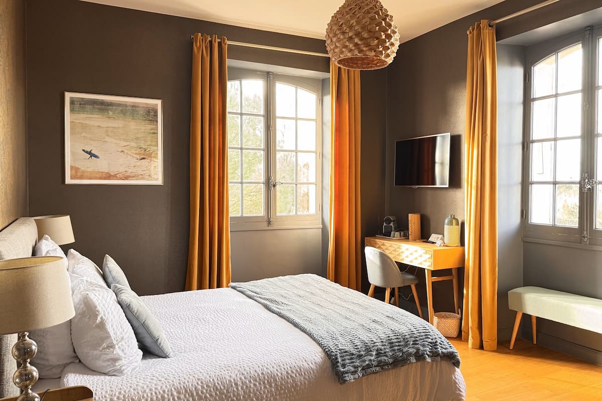 A bedroom with a bed, a desk, a chair, a TV, and large windows with curtains.