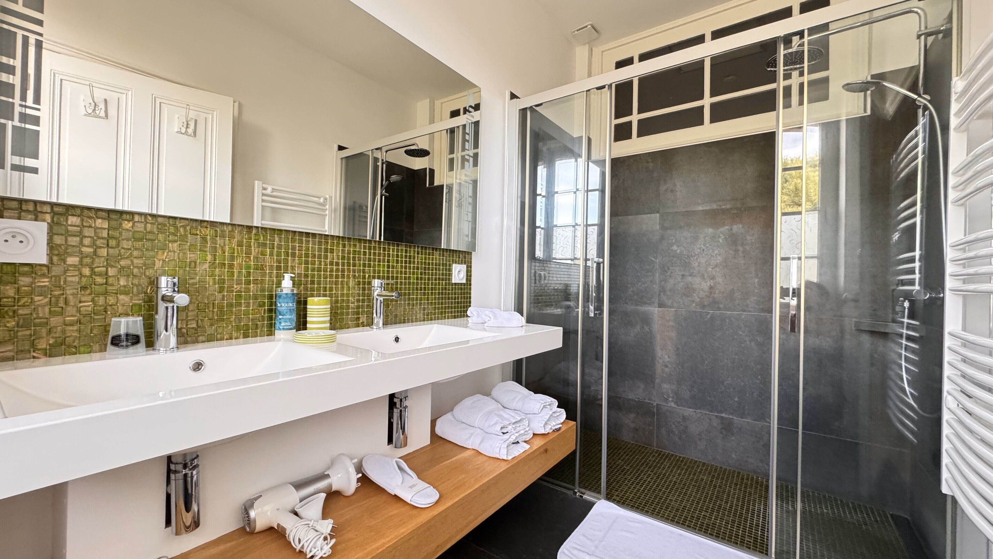 Une salle de bain moderne avec un grand miroir, deux lavabos, une douche et une étagère en bois où sont posées des serviettes.