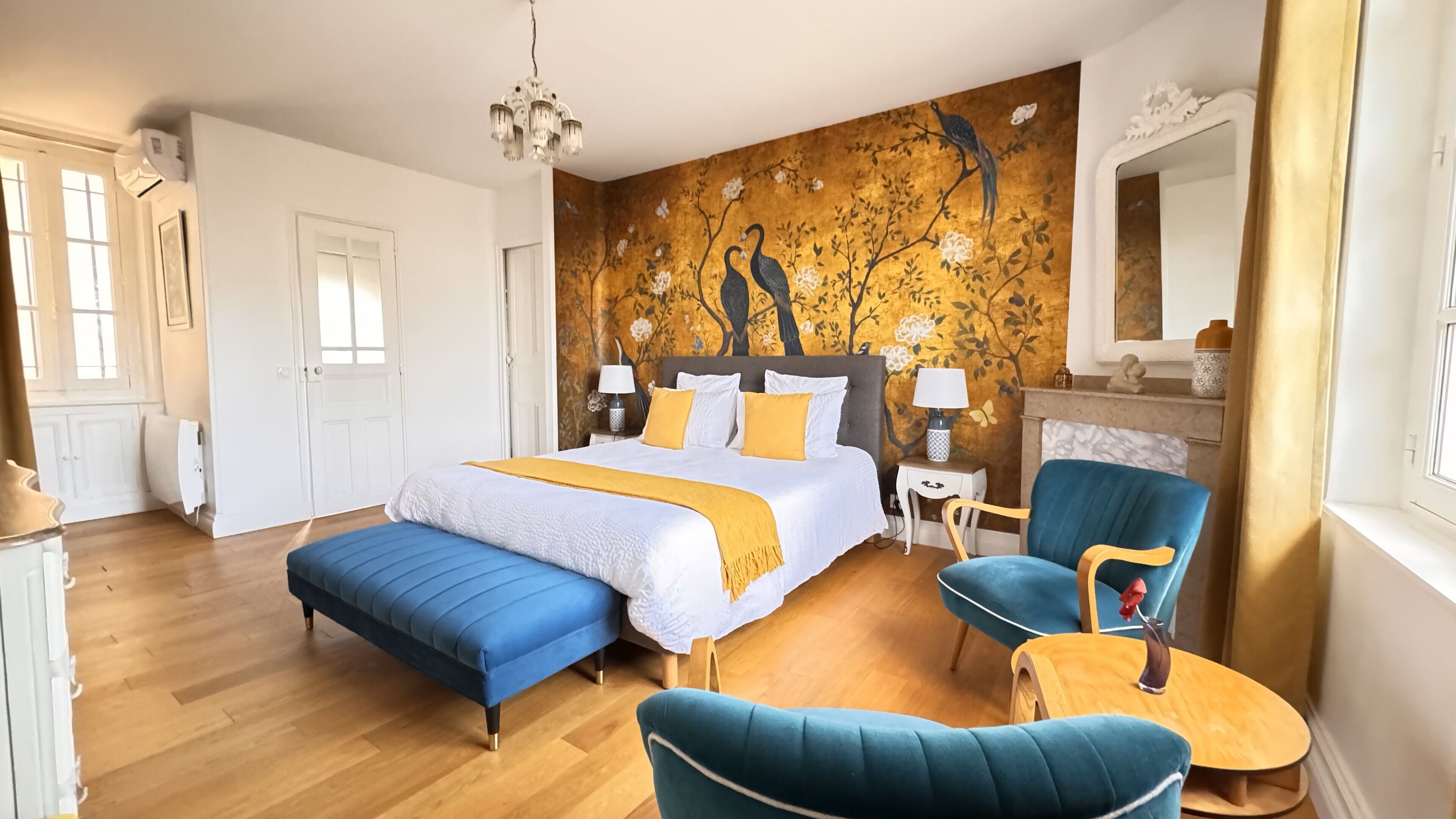 A bedroom with a large bed, two chairs, a small table, a nightstand, and a wall mural.