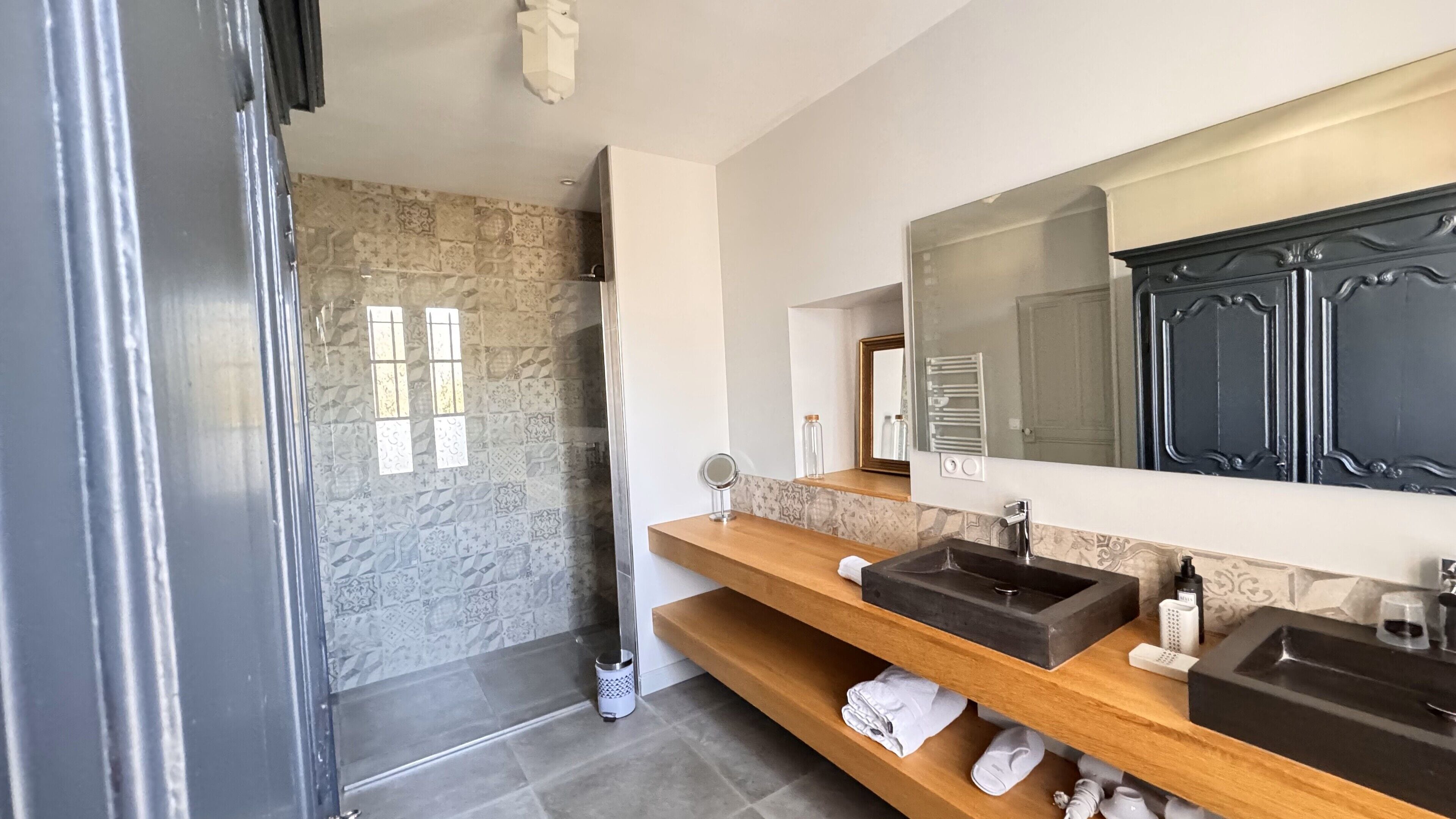 A modern bathroom with a stone shower, two sinks, a large mirror, and a wooden vanity.