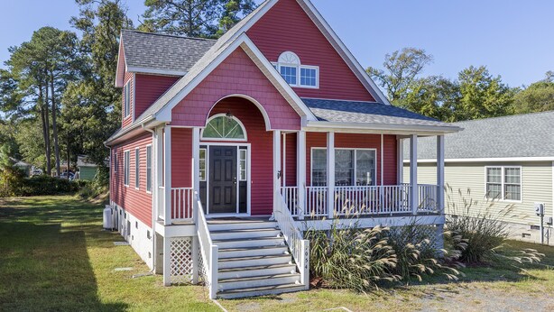 Une maison à deux étages, avec une façade rouge, des éléments décoratifs blancs et un porche à l’avant, accessible par quelques marches menant à une porte.