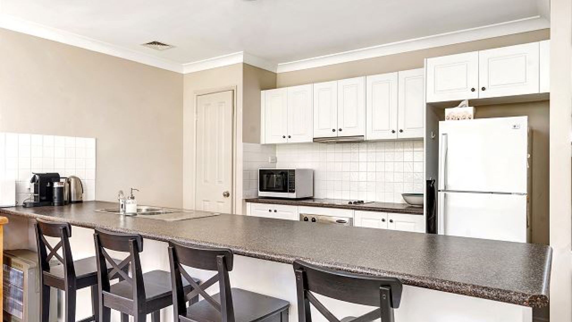 A modern kitchen with white cabinets, a microwave, a refrigerator, and a bar area with chairs.