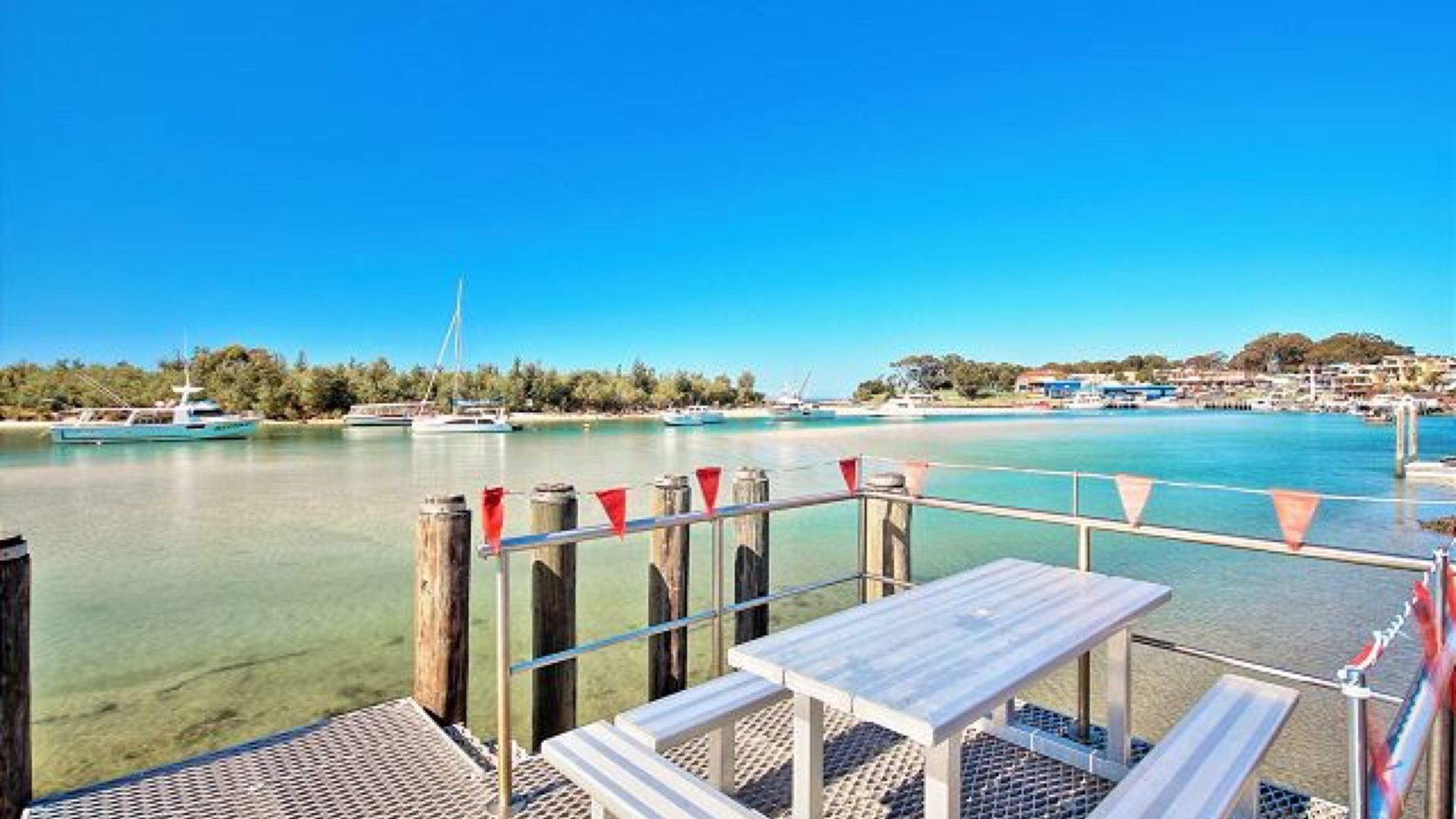 Terraza frente al mar con una mesa y bancos, con vistas a una bahía tranquila con barcos y una costa lejana.
