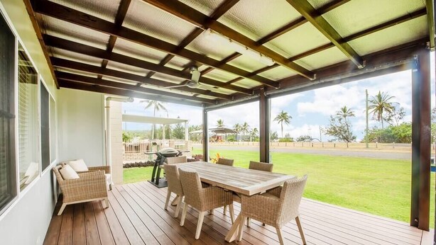 A covered outdoor area with a wooden deck, wicker furniture, and a view of a road and palm trees.