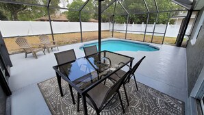 Un patio couvert avec une table et des chaises en verre, une piscine et une vue sur une cour clôturée.