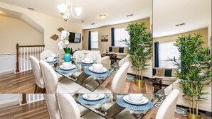A dining area with a glass table, white chairs, and blue placemats. There are two potted plants, a TV on the wall, and a staircase leading to another room.
