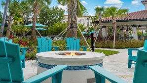 A circular fire pit surrounded by turquoise chairs, with palm trees and a covered seating area in the background.