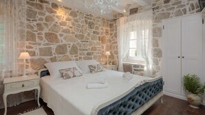 Un dormitorio con pared de piedra, cama con cabecera capitonada, cortinas blancas, candelabro y armario blanco.