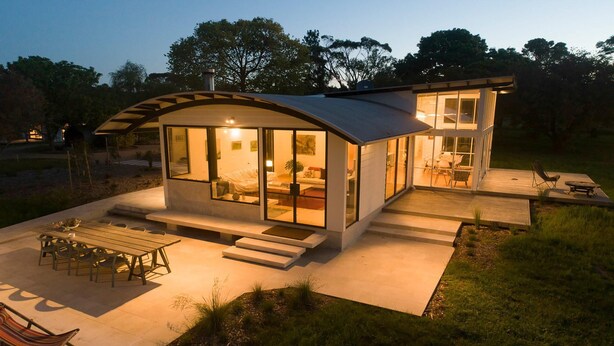 A modern house with a curved roof, large glass windows, and a patio area with outdoor furniture.
