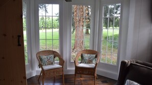 A room with two wicker chairs with patterned cushions, a wooden floor, and large windows with a view of a lawn and trees.