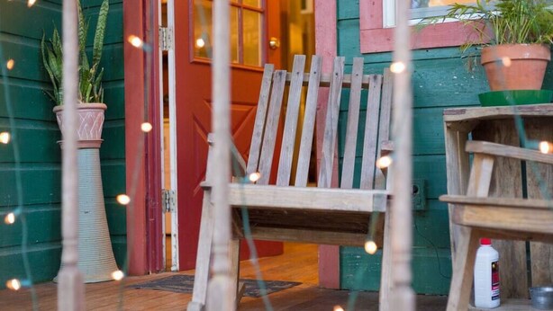 A wooden chair in front of a door with a red frame, a potted plant to the left, and a small table with a bottle on it to the right. - The Victorian Cottage at Hamilton Creek (Burnet County)