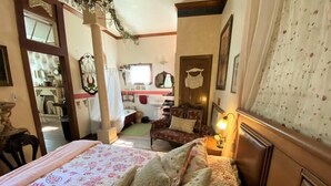 A bedroom with a bed, bedside table, and a chair. There is a window with curtains, a door, and a mirror on the wall. - The Victorian Cottage at Hamilton Creek (Burnet County)