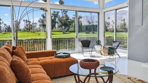 Un salon moderne avec une grande table en verre, un canapé marron et une vue sur un terrain de golf, visible à travers de grandes fenêtres.