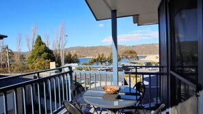 A balcony with a table and chairs, overlooking a parking area and a body of water with hills in the background. - Silvertrees 5 - Central 3BR Townhouse l Lake Views l Spa l BBQ l WiFi (Jindabyne)