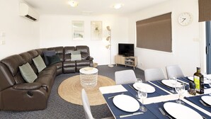 Ein modernes Wohnzimmer mit einem braunen Ledersofa, einem Esstisch für vier Personen, einem Flachbildfernseher und einem großen Fenster mit Jalousien.