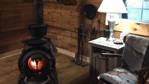 A cozy room with a wood-burning stove, a wooden chair, a table with a lamp, and a window with curtains.