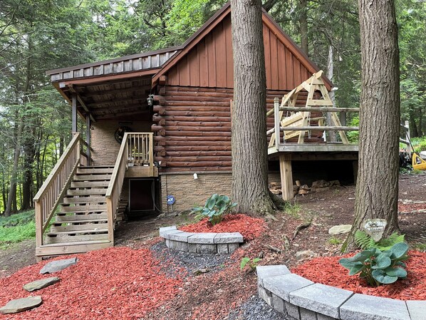 A log cabin with a wooden staircase leading to a deck, surrounded by trees and a red mulch garden.